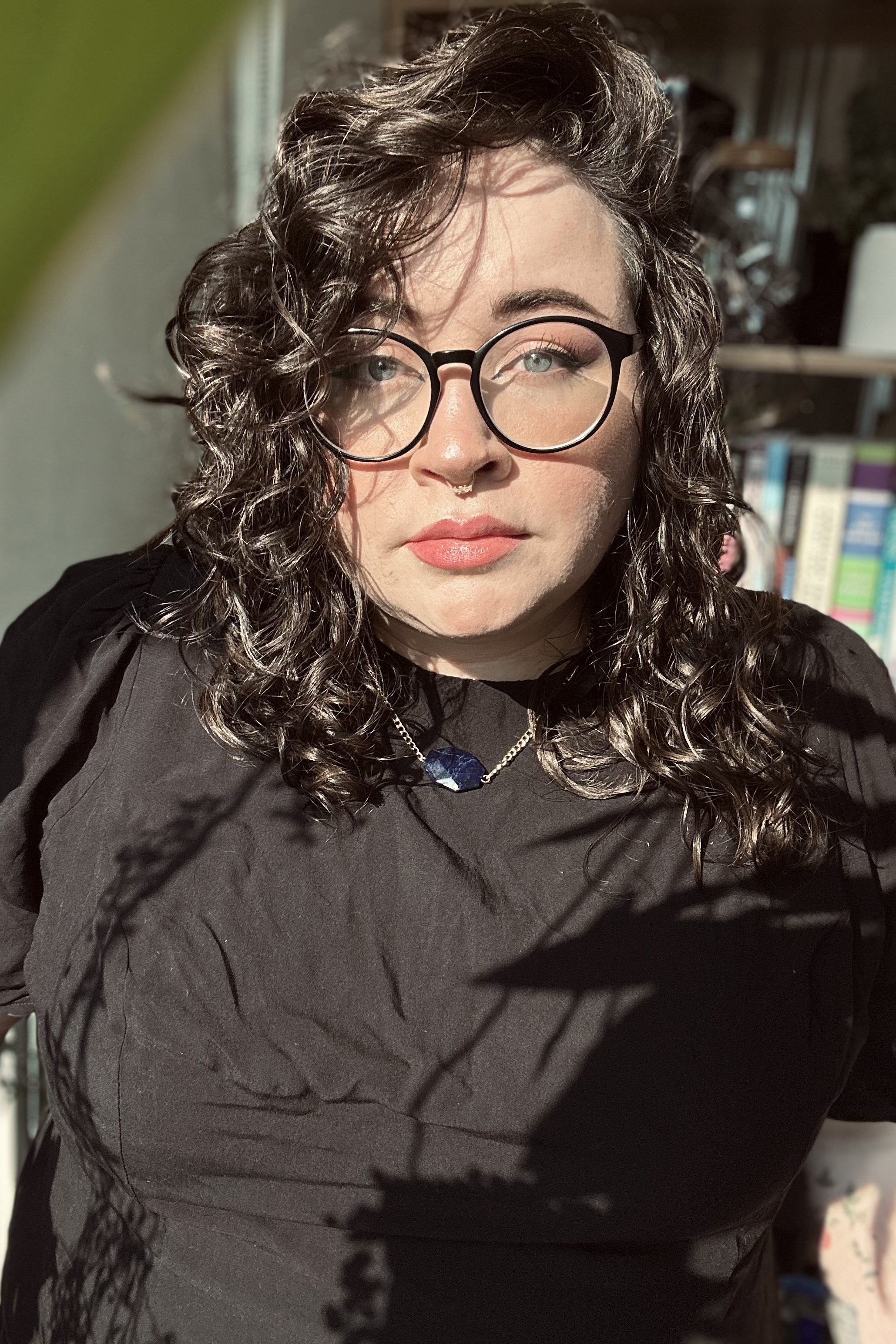 hannah standing in dappled sunlight, their hair curly with a soft expression.